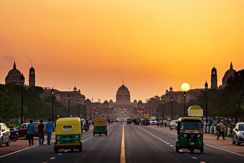 A wide shot of a bustling Delhi street during a hot summer day, with clear blue skies. People are seen seeking shade under awnings or trees, some covering their heads with scarves, and a few vehicles are visible. The atmosphere appears hazy due to heat.