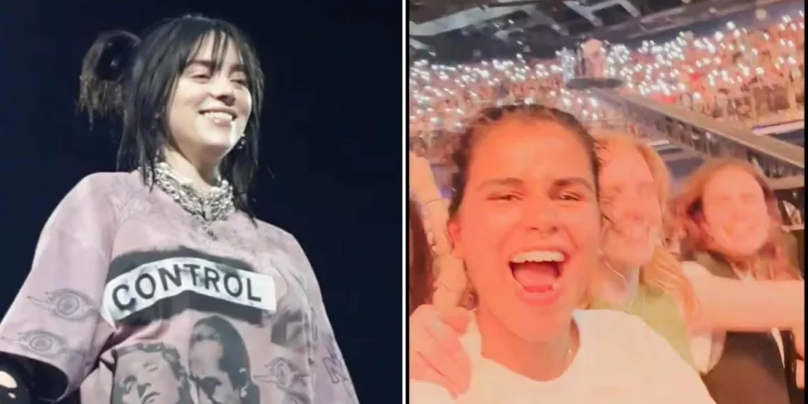 Billie Eilish smiling on stage, wearing a baggy outfit, with bright lights behind her.
