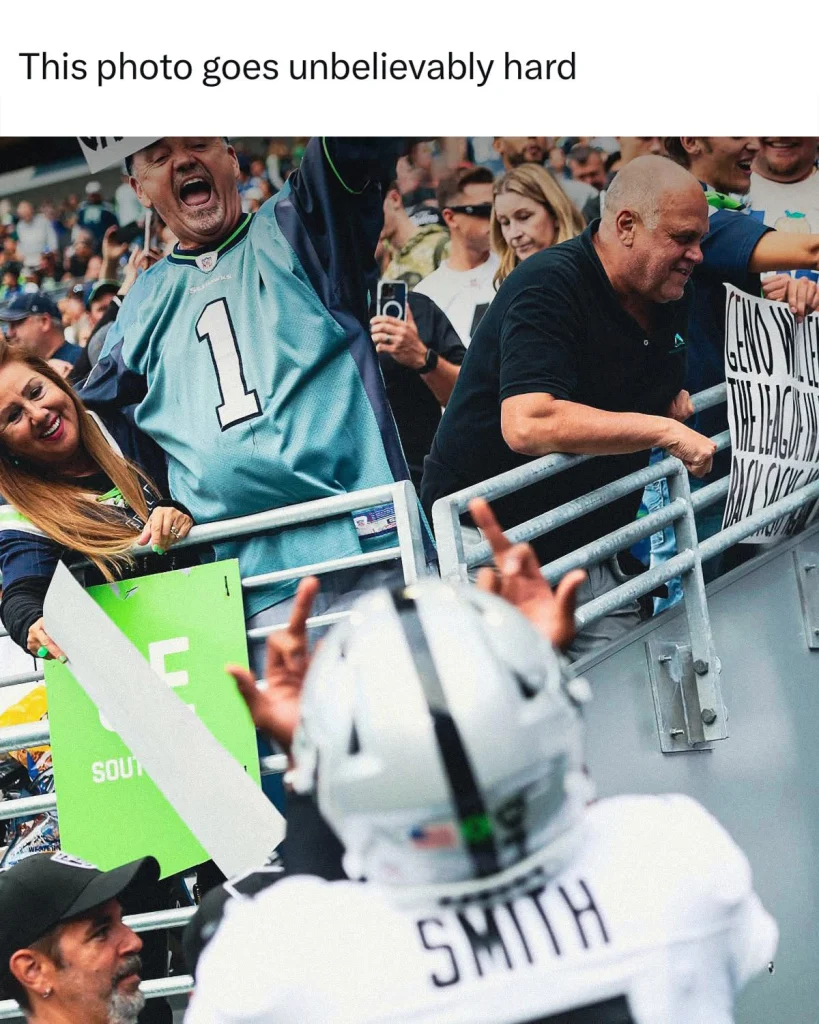 fan photo capturing football player Geno Smith in a white and silver jersey with “SMITH” on the back, facing away from the camera. He raises his hands toward an excited crowd behind a railing, including a man in a Seahawks jersey yelling and another holding a sign. The caption reads, “This photo goes unbelievably hard.”