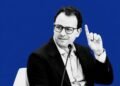 Black and white portrait of Anthropic CEO Dario Amodei raising a finger during a panel discussion, set against a solid bold blue background.
