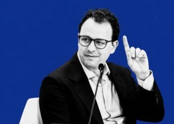 Black and white portrait of Anthropic CEO Dario Amodei raising a finger during a panel discussion, set against a solid bold blue background.