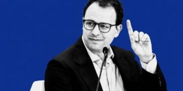 Black and white portrait of Anthropic CEO Dario Amodei raising a finger during a panel discussion, set against a solid bold blue background.