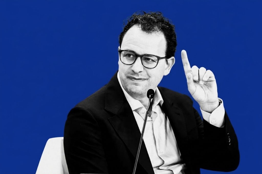 Black and white portrait of Anthropic CEO Dario Amodei raising a finger during a panel discussion, set against a solid bold blue background.